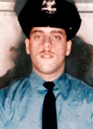 Edward Byrne smiling, wearing a black police cap, blue long sleeves, and a black necktie.
