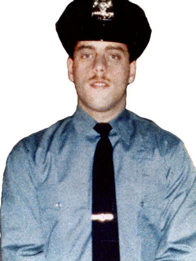 Edward Byrne smiling, wearing a black police cap, blue long sleeves, and a black necktie.
