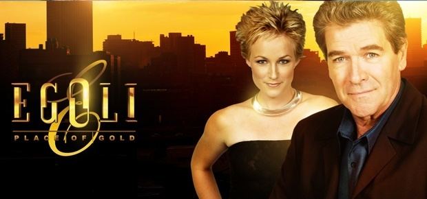 Lindie Van Eeden and Eckard Rabe are smiling while Lindie is wearing a black top and Eckard is wearing a black coat and long sleeves in the 1992 soap opera, Egoli: Place of Gold