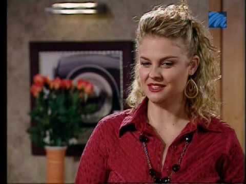 A woman with a blonde curly hair is wearing a red blouse and necklace in a scene from the 1992 soap opera, Egoli: Place of Gold
