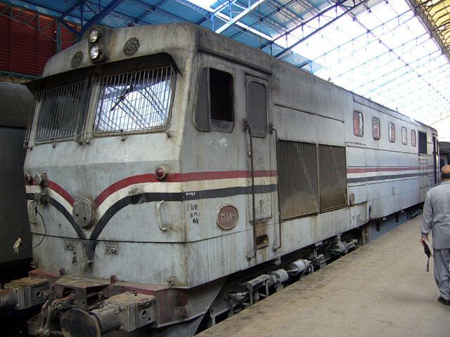 Egyptian National Railways, It has train railways, and a single old gray train wagon parked at the station at the right is a man standing, back view holding a small bag with a bald top and black hair wearing a gray long sleeve shirt and gray pants.