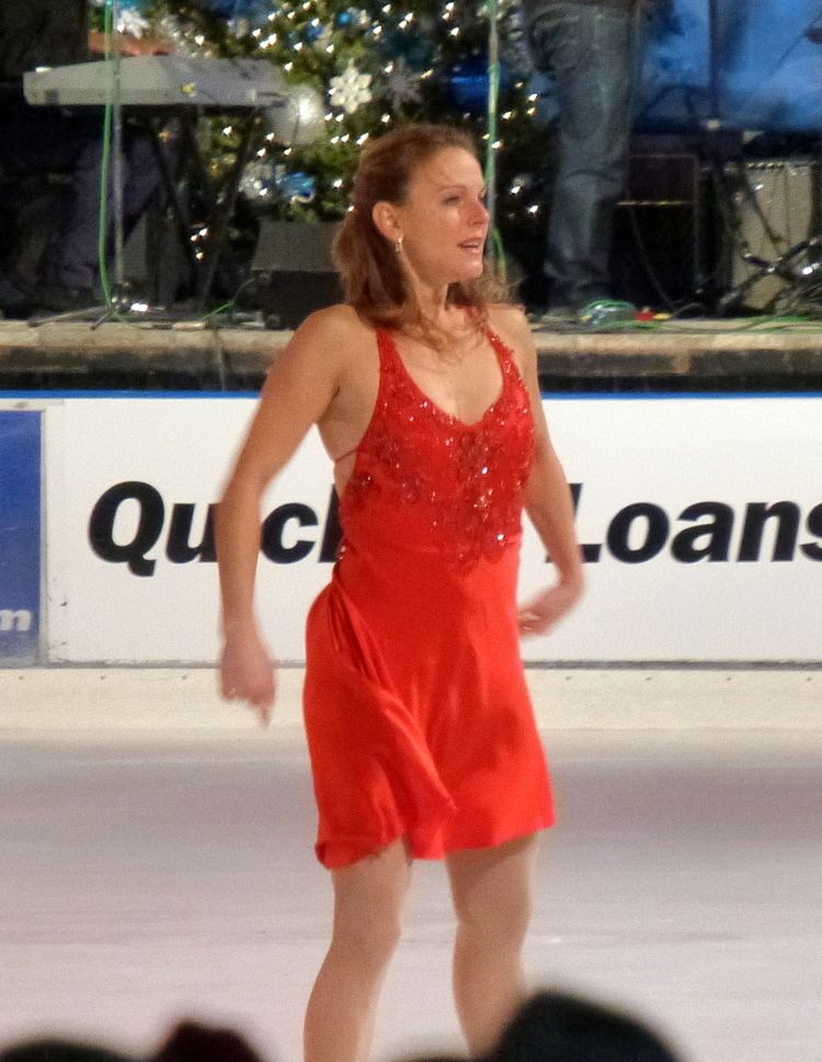 Ekaterina Gordeeva smiling, looking sideways, a Russian figure skater, performing at the 2014 Detroit Christmas Tree Lighting Ceremony at Campus Martius Park, with a band and Christmas decorations in the background. She has short blonde hair half-tied flowing, has earrings on, wearing a deep-V, bareback with ties in a red beaded figure skating dress with a visible cleavage.
