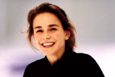 Ekaterina Gordeeva smiling, a Russian figure skater with messy brown hair tied, thin loop earrings, wearing a black knitted neck cut sweater.