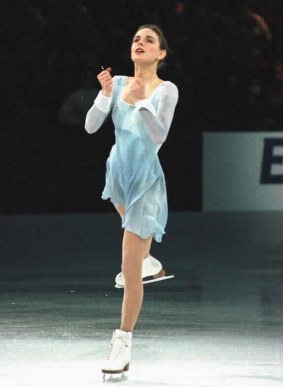 Ekaterina Gordeeva looking up afar, closed fists while doing a figure skating stunt, is a Russian figure skater. Her hair is neatly bun, wearing a pair of white figure skates, and a pale blue collared sleeves figure skating dress.