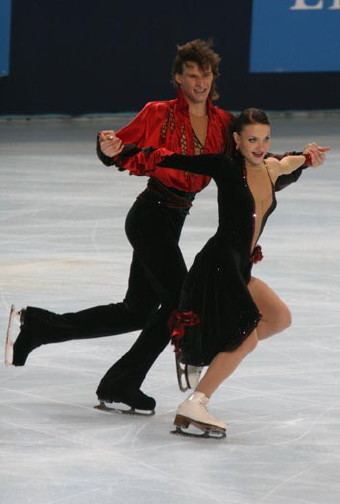 Ekaterina Rubleva and Ivan Shefer are smiling while performing ice dancing. Ekaterina is wearing earrings, a black dress, and white skate boots while Ivan is wearing red long sleeves, black pants, and black skate boots.