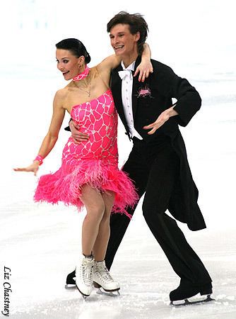 Ekaterina Rubleva and Ivan Shefer are smiling while performing ice dancing. Ekaterina is wearing earrings, a necklace, a pink choker, a pink dress, and white skate boots while Ivan is wearing a black coat over white long sleeves, a white bowtie, black pants, and black skate boots.