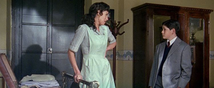 In the movie scene of El Palomo cojo, María Barranco (left) is serious, looking at the Miguel Ángel Muñoz while talking, her right hand holding the maid's trolley and left hand on her waist, has brown curly hair wearing a maids outfit, on the right is Miguel Ángel Muñoz standing, listening to the maid while his right hand down while his left arm is in the pocket and a cabinet behind him, has brown hair wearing a black shirt over white polo and a maroon necktie and gray coat.