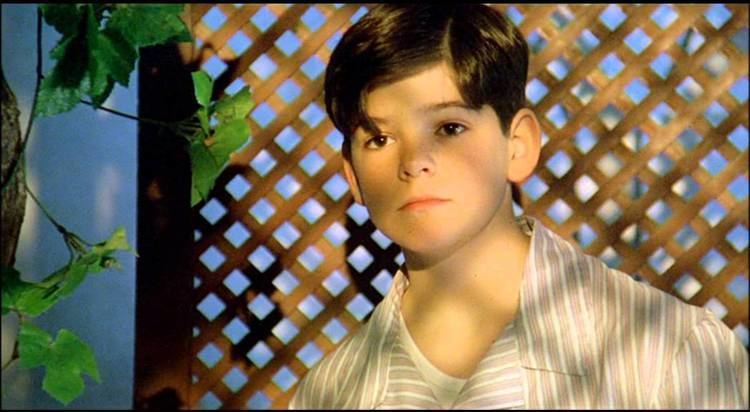 In the movie scene of El Palomo cojo 1995, Miguel Ángel Muñoz  is serious, standing on the right of leafy vines and trellis at the back. Has brown hair wearing a gray striped polo.