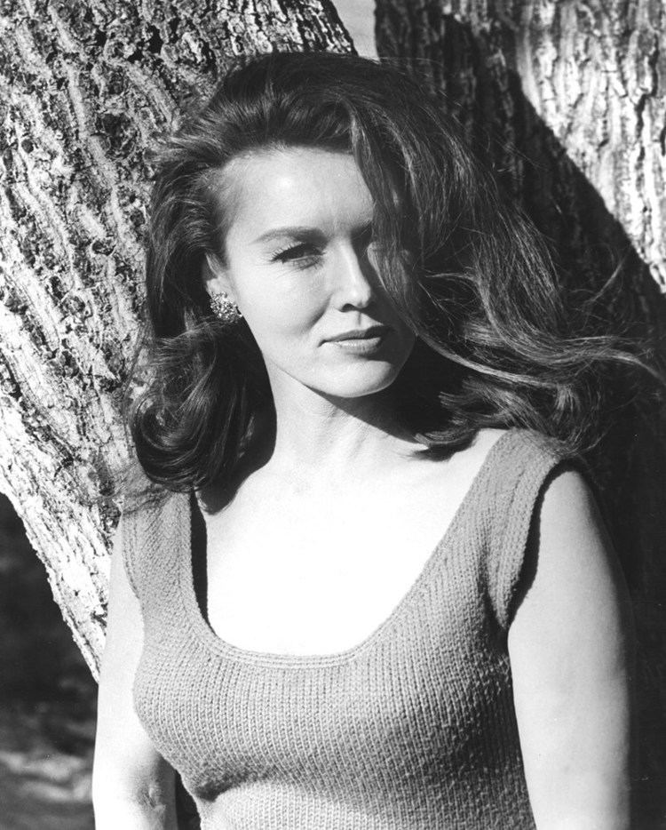 Elaine Devry with wavy hair, wearing earrings and sleeveless top while leaning on a tree.