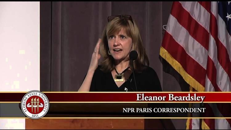 Eleanor Beardsley giving a speech at Florida State University with a serious face and behind her is the Flag of the United States. Eleanor is wearing a necklace, earrings, a ring, eyeglasses on the top of her head, and a black blouse