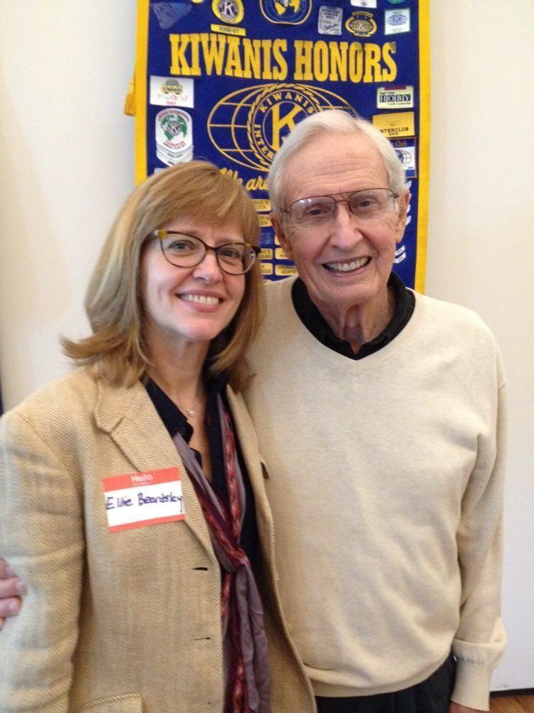 Eleanor Beardsley and the man beside her are smiling and behind them is a board with Kiwanis honors' logos. Eleanor is wearing eyeglasses, a necklace, a purple and red ribbon scarf, and a black blouse under a beige coat with a name tag on the coat. The man beside her is wearing eyeglasses and a black shirt under a cream sweatshirt