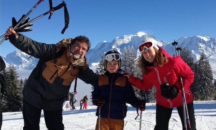 Ulysse Gosset smiling with his son, Maxime, and wife, Eleanor Beardsley while holding ski poles during their ski vacation in the Alps, with trees and mountains in the background. Ulysse with a mustache and beard is wearing eyeglasses, a black and brown jacket, and black pants. Maxime is wearing black gloves, goggles, a black and brown jacket, and brown pants. Eleanor is wearing black gloves, goggles, a red jacket, and black pants