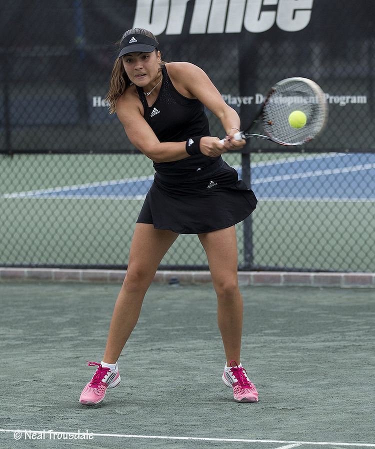 Elena Gabriela Ruse playing tennis while wearing a black top, black skirt, black cap, and pink rubber shoes