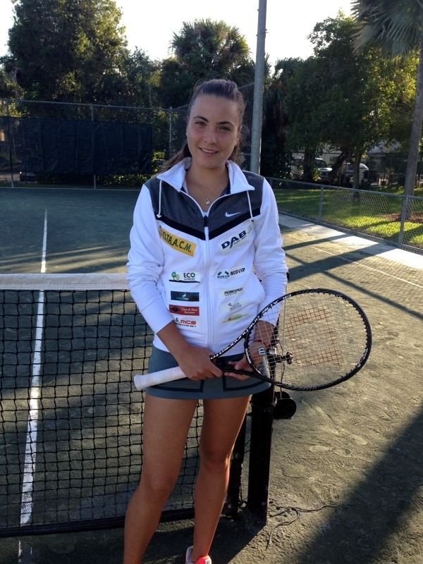 Elena Gabriela Ruse smiling while holding a racket and wearing a black and white jacket and gray skirt