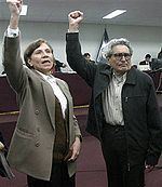 Elena Iparraguirre and Abimael Guzmán raising their hands while she is wearing a beige blazer, brown vest, and white blouse