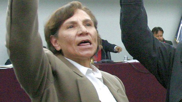 Elena Iparraguirre raising her hand while wearing a beige blazer, brown vest, and white blouse