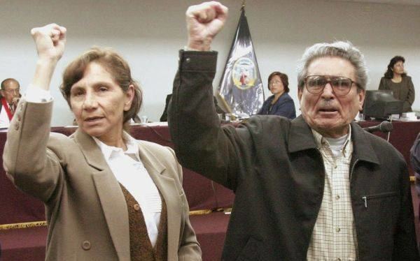 Elena Iparraguirre and Abimael Guzmán raising their hands while she is wearing a beige blazer, brown vest, and white blouse
