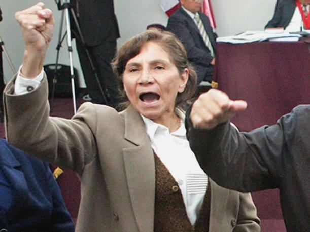 Elena Iparraguirre raising her hand while wearing a beige blazer, brown vest, and white blouse