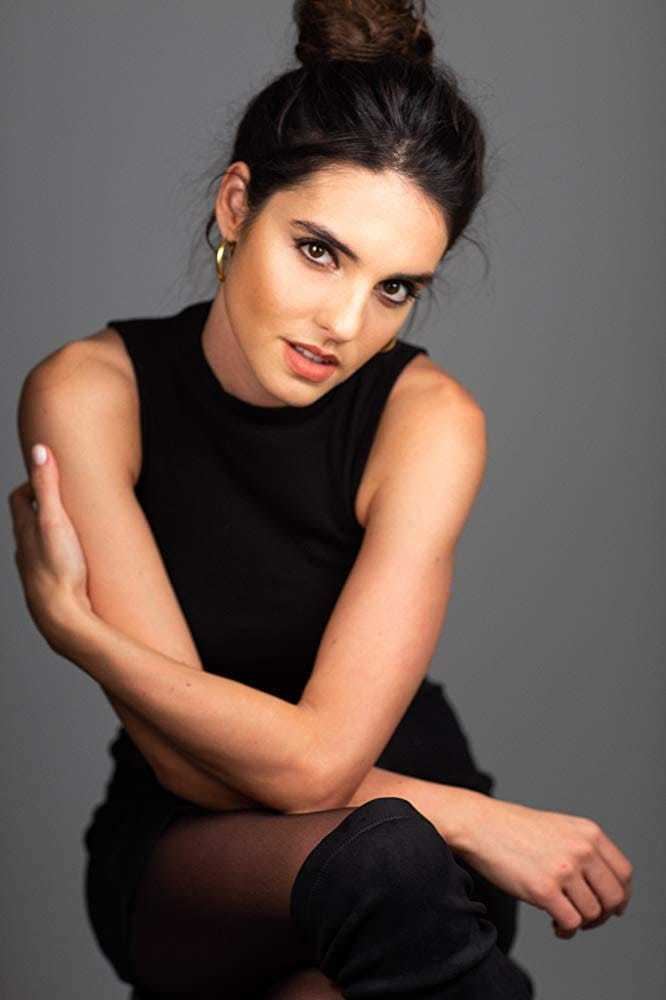 Elena Rusconi smiling while sitting with her hair in a bun style and wearing a black tank top, stockings and gold round earrings