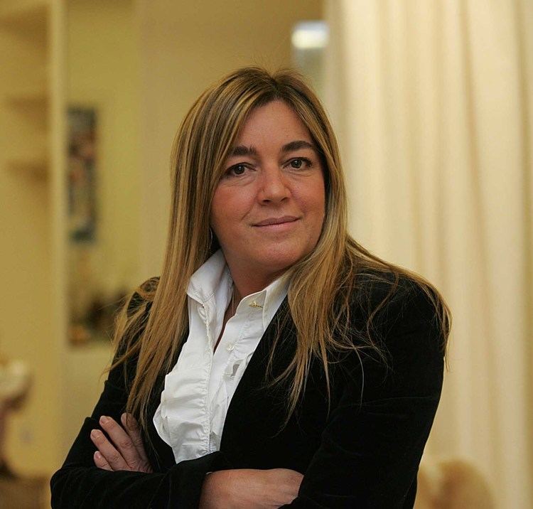 Eliana Tranchesi is smiling while standing and hand crossed, has long dark blond hair wearing white blouse and black coat.