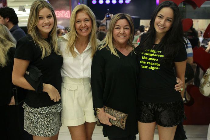 From left Marcella Tranchesi (1st) is smiling, standing while holding her bag with her arm, has long blond hair wearing black shirt and animal printed skirt, next to her is a woman standing while holding their hand at the back has long blonde hair wearing white blouse and white shorts, Eliana Tranchesi (3rd) is standing posing with straight body while holding her animal printed bag on her right hand has dark blond hair wearing black v cut formal dress, Luciana Tranchesi (4th) is standing beside her mom (3rd) has black long hair wearing black printed shirt and black shorts.