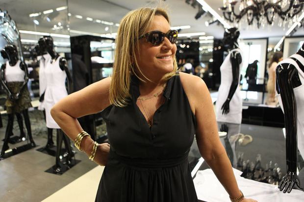 Eliana Tranchesi is smiling, posing with the right hand on her waist and left hand on the table, behind her back are black mannequins wearing different kind of outfits, she has long blond hair wearing shades, gold bracelets and black dress.