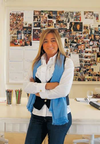 Eliana Tranchesi is smiling, standing arms crossed, behind is a wall board with camera photos and papers stick on it, a white table with two cups full of colored pencils on her right and an open notebook with black pen on top and wine glass with water on her left, She has long dark blond hair wearing round earrings, a white blouse, denim pants, and a blue scarf over a black vest.
