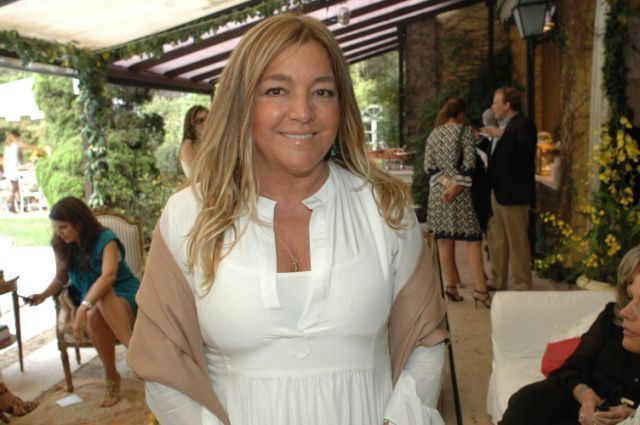 Eliana Tranchesi is smiling while standing in front of people has long blond hair wearing white dress, necklace and a brown cloth.