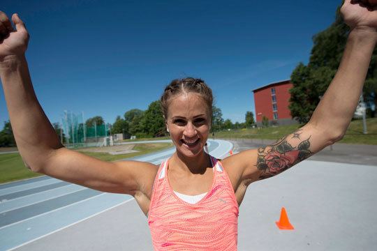 Elin Westerlund smiling beside the running track with tattoos on her left arm, raising both of her hands and wearing a white and orange shirt