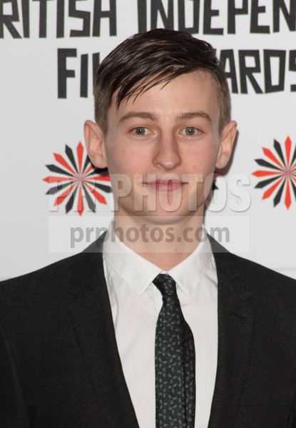 Elliott Tittensor smiling, wearing a black coat over white long sleeves and a black tie.