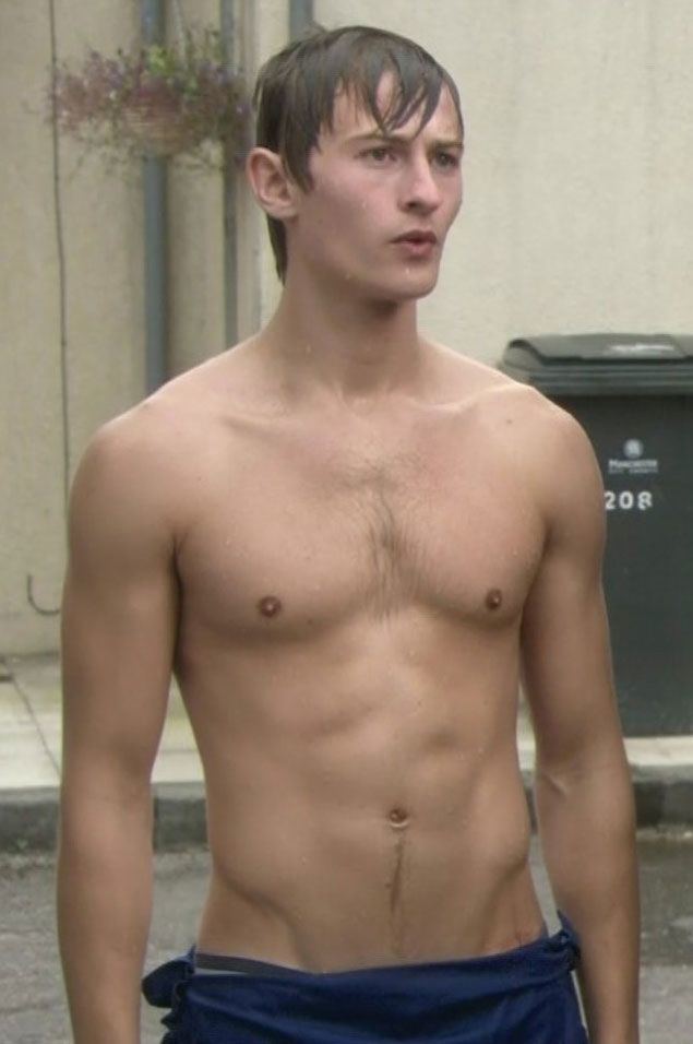Elliott Tittensor with a curious face while topless, with wet body and hair, and wearing blue shorts.