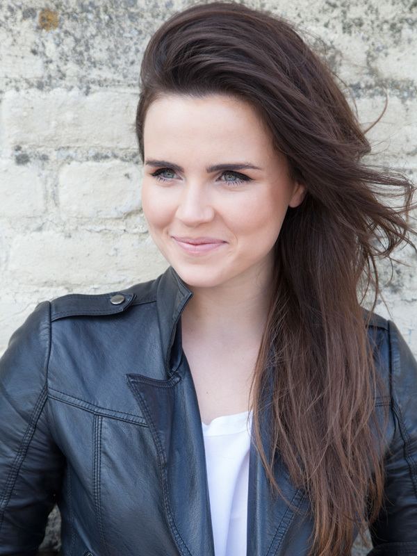 Emer Kenny is smiling while looking at something and leaning on the wall, with long black hair, and wearing a black leather jacket over a white top.