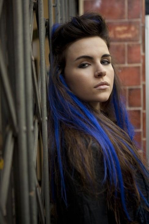 Emer Kenny with a serious face while leaning on a steel gate, with black and blue color hair, and wearing a black long sleeve top.