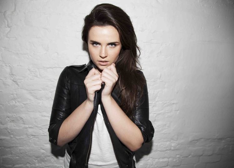 Emer Kenny with a serious face while holding her black leather jacket, with long black hair and wearing a white shirt.