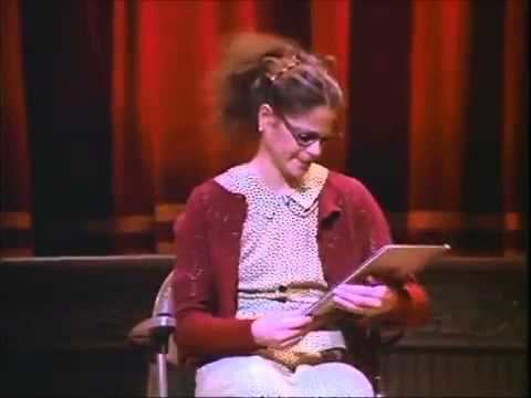 Emily Litella looking at the paper while wearing a red crochet blazer, polka dot blouse, and eyeglasses at the Saturday Night Live