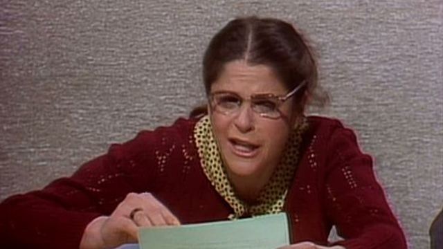 Gilda Radner as Emily Litella talking to someone while wearing a red crochet blazer, polka dot blouse, and eyeglasses