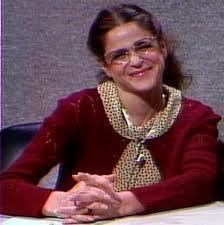 Gilda Radner as Emily Litella smiling while wearing a red crochet blazer, polka dot blouse, and eyeglasses at the Saturday Night Live
