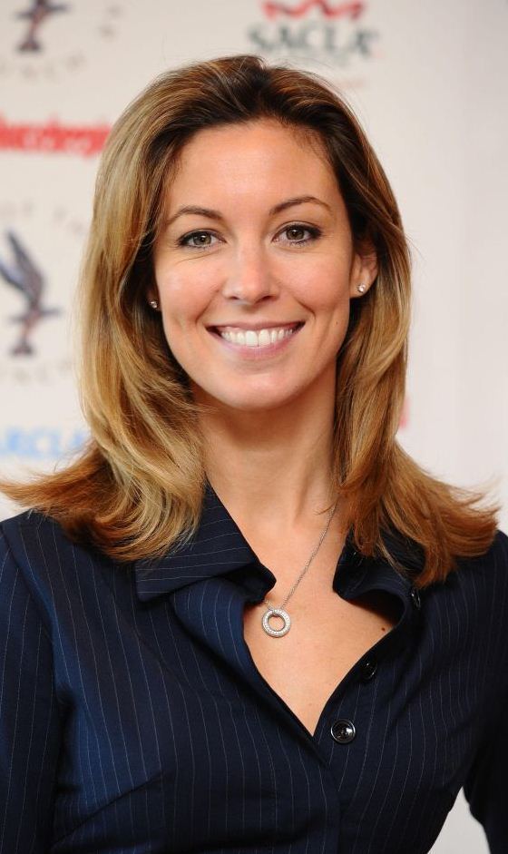 Emma Crosby smiling, with blonde hair, wearing earrings, a necklace, and a striped black top.