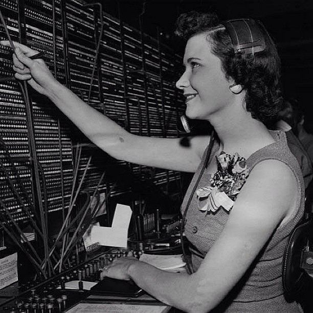 A younger Emma Nutt smiling while operating some telephone lines with a corsage on her clothing.