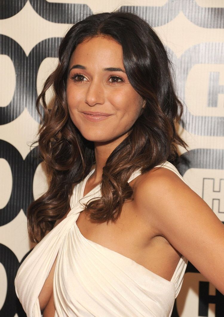 Emmanuelle Chriqui smiling while looking on a side with her wavy hair down and wearing a wrap cross-tied backless white dress