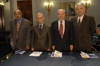 The members of the 2007 inductees- Emmett Chappelle, John Franz, Paul Baran, and Robert Metcalfe (from left to right). Paul is smiling while Emmett, John, and Robert are having serious faces with a table in front of them, with some papers on it, there are lights on the wall, and a painting of a man. Emmett is wearing a blue long sleeve under a yellow and blue necktie and a gray coat. John is wearing a gray long sleeve under a black necktie and brown coat. Paul is wearing a white long sleeve under a red necktie and black coat. Robert is wearing a white long sleeve under a black patterned necktie and brown coat
