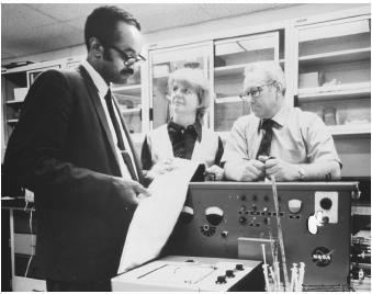 Emmett W. Chappelle (left) holding a long paper, looking at it, and standing beside the machine while the woman (center) and a man (right) are looking at him and there are shelves at the back. Emmett with a mustache and beard is wearing eyeglasses and a long sleeve under a necktie and a coat. The woman (center) is wearing a blouse under a vest while the man (right) is wearing eyeglasses and long sleeves with a necktie