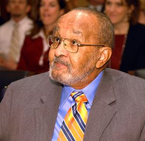 Emmett Chappelle looking at something during the National Inventors Hall of Fame induction ceremony in 2007. Emmett with a serious face, mustache, beard and there are people in the background. Emmett is wearing eyeglasses and a blue long sleeve under a yellow and blue necktie and a gray coat