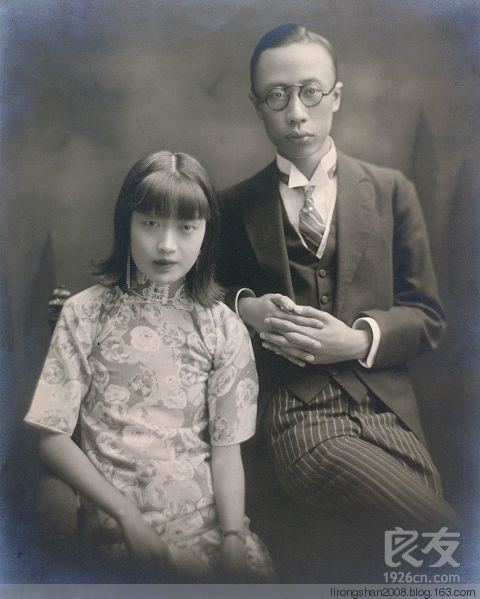 Empress Wanrong and Puyi with serious faces. Empress Wanrong wearing earrings and a qipao dress while Puyi wearing eyeglasses, a black coat, a black vest over a white long sleeve, and black striped pants.