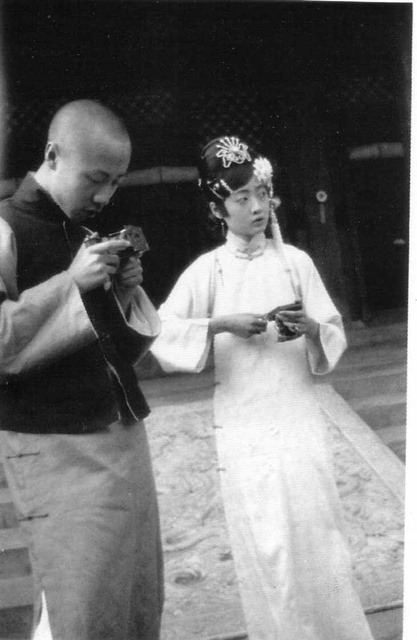 Empress Wanrong with a serious face with a man looking at something on his hand. Empress Wanrong wearing a headdress, earrings, and a white qipao dress.