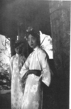 Empress Wanrong with a serious face with a lady on her back. Both are wearing a headdress and a white floral qipao dress.