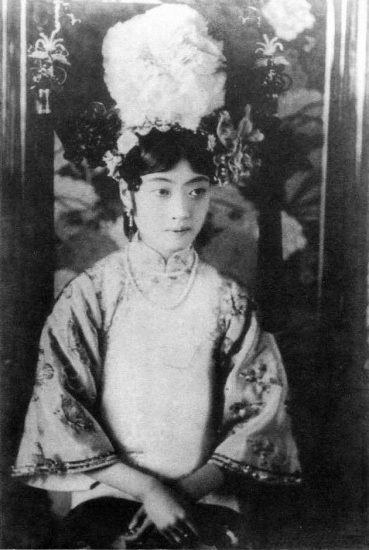 Empress Wanrong with a serious face, wearing a headdress, a necklace, and a qipao dress.