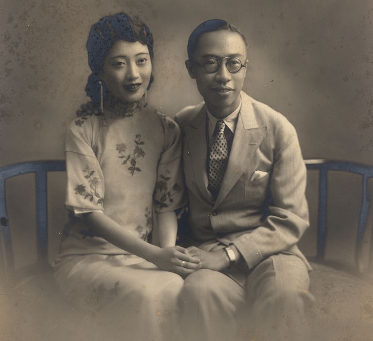 Empress Wanrong and Puyi are smiling while sitting on a chair. Empress Wanrong wearing earrings and a floral qipao dress while Puyi wearing eyeglasses, a coat, a white long sleeve, and a polka dot necktie.