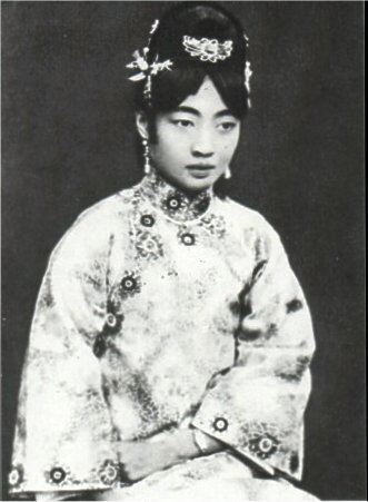 Empress Wanrong with a serious face, wearing a headdress, earrings, and a qipao dress.
