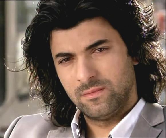 Engin Akyurek with long hair while wearing a gray coat and white long sleeves